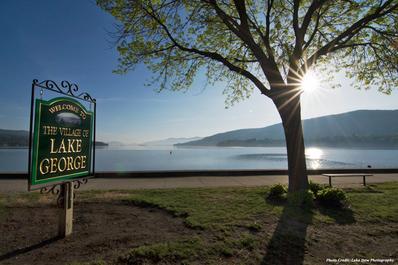 welcome to the village of lake george sign in front of the lake in spring